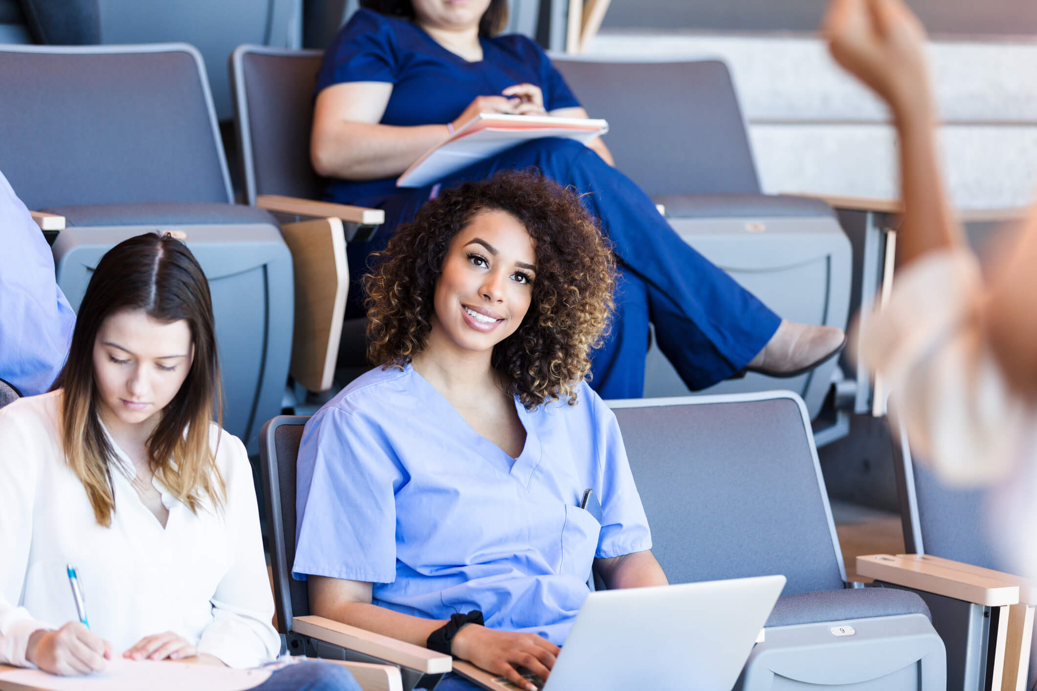 A female African-American nursing student smiles while taking notes on her laptop during a lecture. She is wearing light blue scrubs. Her classmates are sitting next to her and in the auditorium rows in the background.